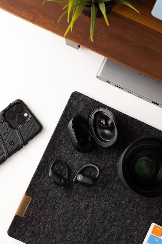 ZIZO Encore Sport Active Wireless Earbuds in Black resting on a dark desk mat beside a phone and a lens