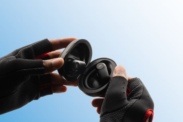 ZIZO Encore Sport Active Wireless Earbuds in Black in their open charging case, held by gloved hands against a blue sky