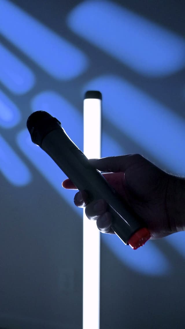 A hand holding the ZIZO Boom's wireless microphone beside a lit tube light in a dark, blue-lit room