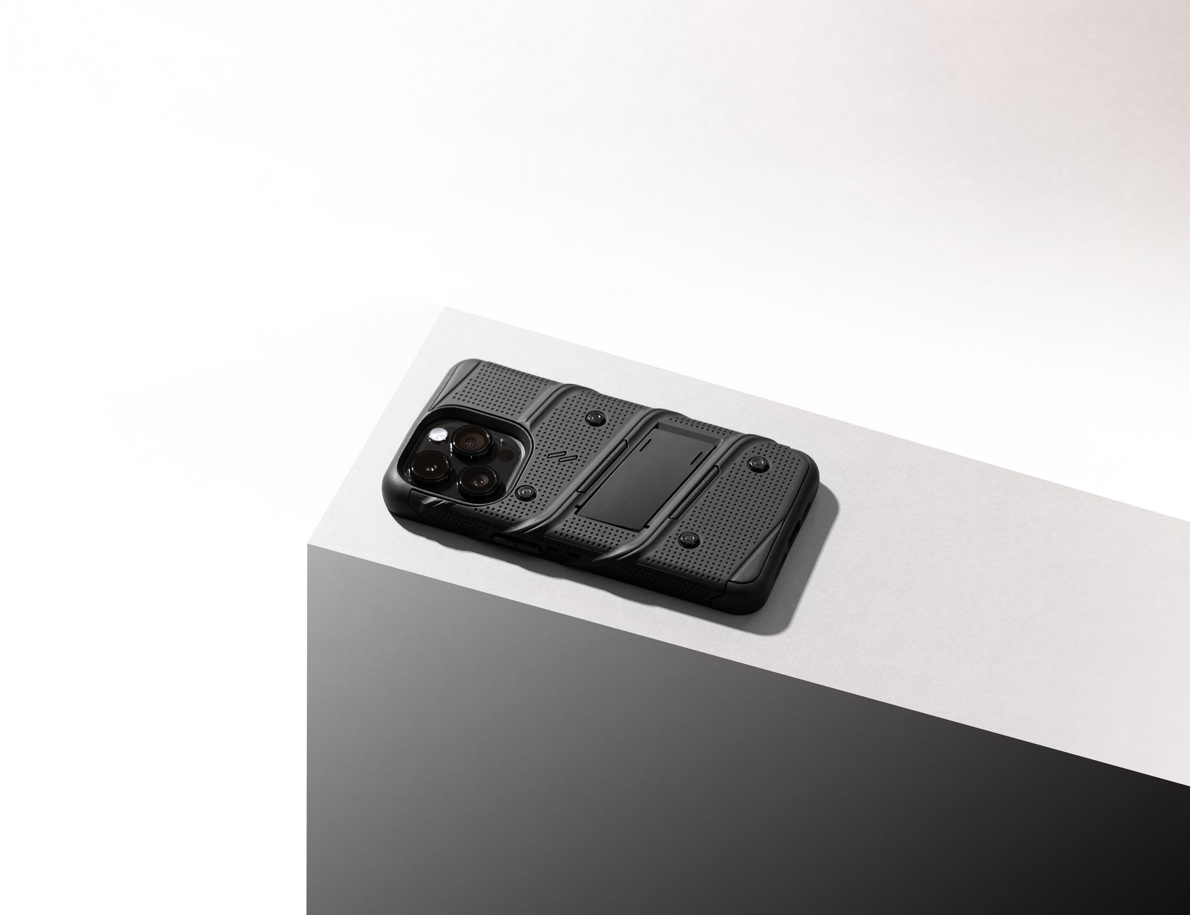 Black ZIZO Bolt rugged case with dot-grid textured back and built-in kickstand, on an iPhone, shot at an angle on a white pedestal edge